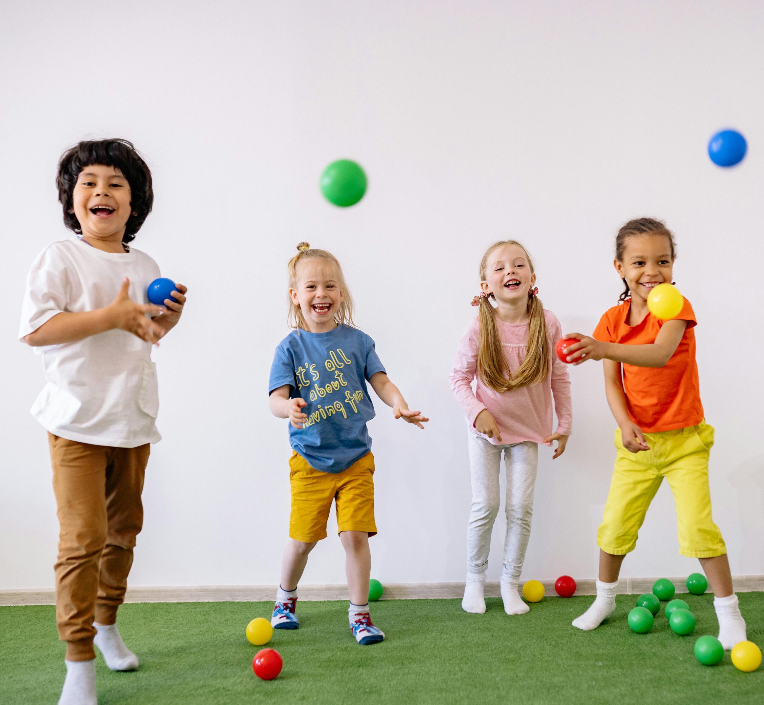 Four diverse children having fun with colorful balls in a bright indoor setting.